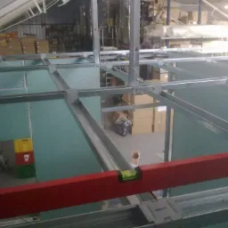 Montaż sufitu podwieszanego z płyt kartonowo-gipsowych w magazynie, widoczna stalowa konstrukcja nośna i poziomica w kolorze czerwonym.