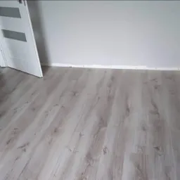 Jasne panele podłogowe ułożone w pokoju z białą ścianą i drzwiami z przeszkleniami; widoczne ślady silikonu przy listwie przypodłogowej.