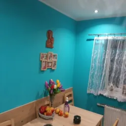 Turkusowa ściana w jadalni po malowaniu, z drewnianą boazerią, dekoracjami ściennymi i stołem z owocami i kwiatami.