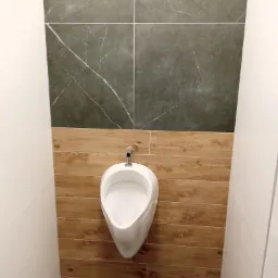 Wykończona toaleta publiczna w Nowych Skalmierzycach z białym pisuarze na ścianie wyłożonej panelami imitującymi drewno, nad którym znajdują się szare płytki imitujące marmur.