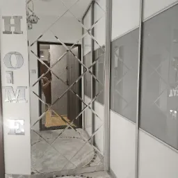 Nowoczesny przedpokój z lustrzaną ścianą w romby i przesuwną szafą w odcieniach szarości i bieli, uzupełniony dekoracją ścienną HOME i okrągłym dywanem.