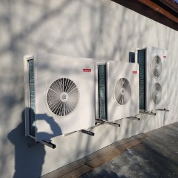 MAT-BUD Mateusz Kaźmierski - Trzy białe jednostki zewnętrzne klimatyzacji Ariston zamontowane na ścianie budynku, rzucające cień drzewa. Widoczne wsporniki montażowe i fragment dachu.