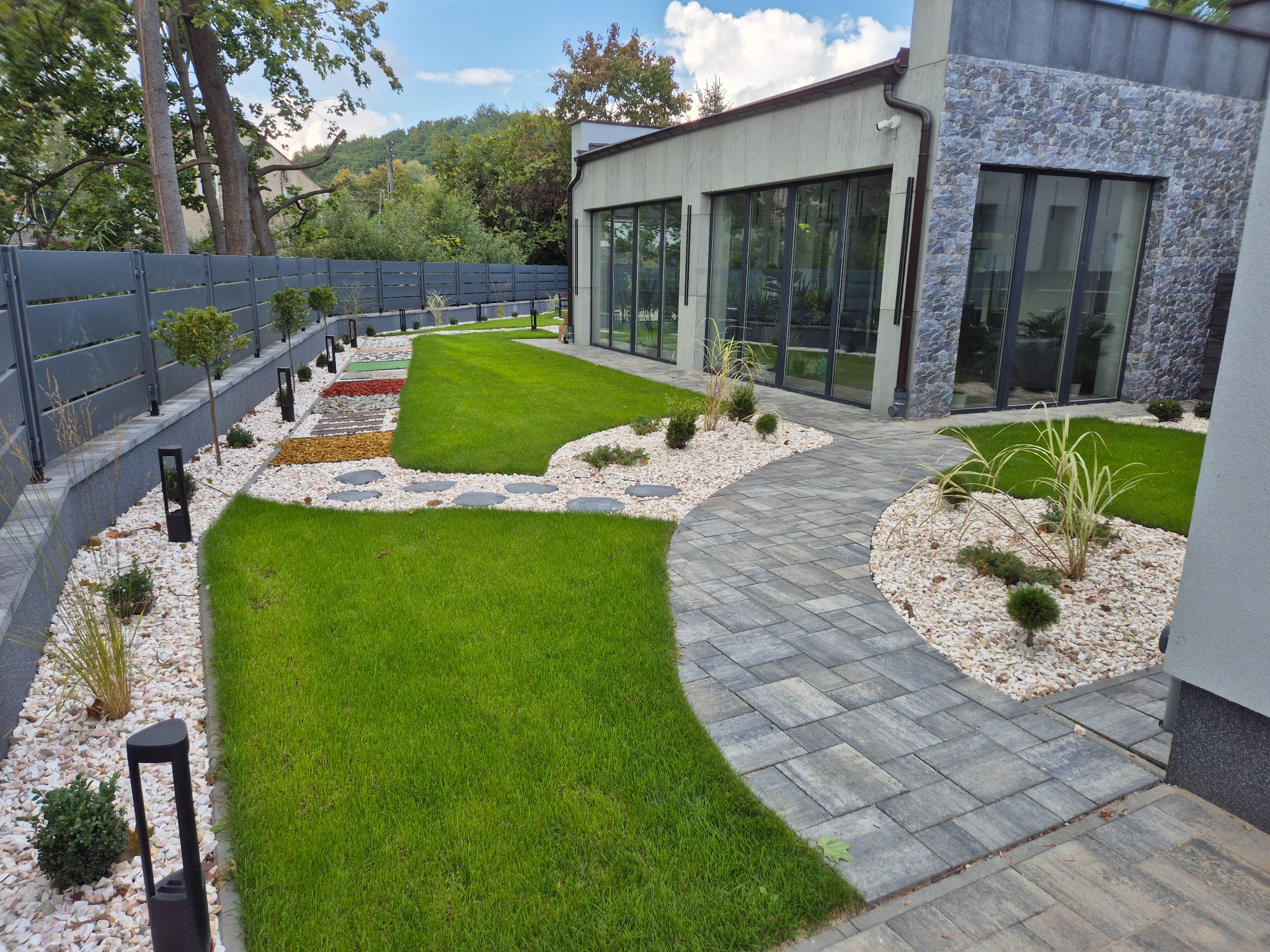 Nowoczesny ogród z równo przystrzyżonym trawnikiem, ścieżką z kostki brukowej i rabatami z białego kamienia. Widoczne lampy ogrodowe i szary płot.