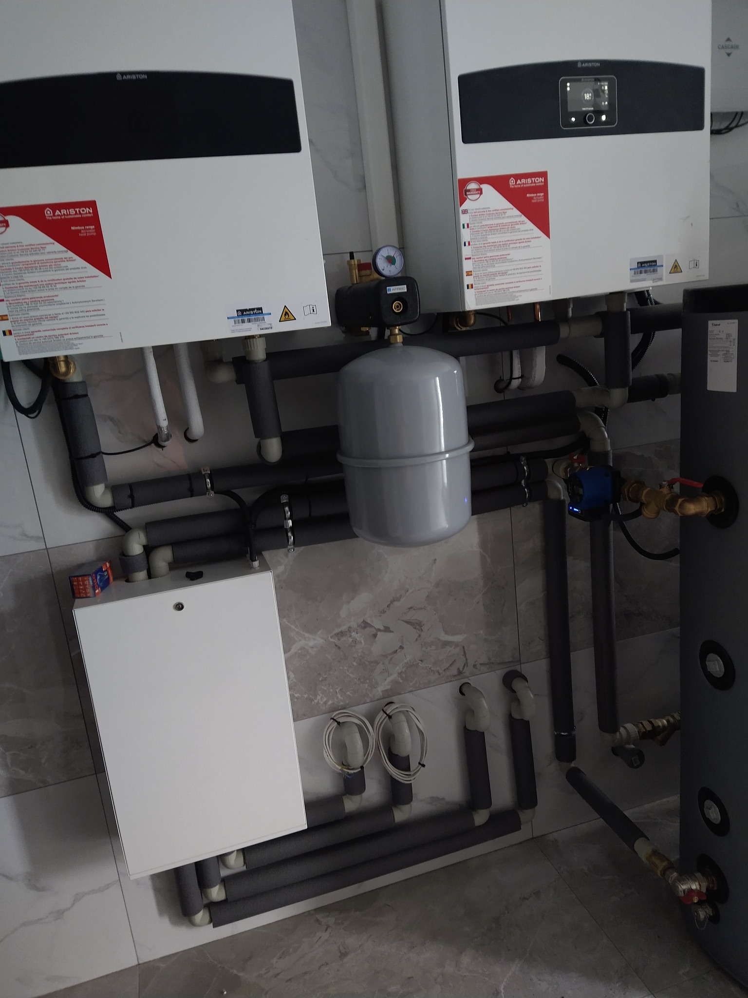 Dwa białe piece gazowe Ariston z szarymi rurami i zbiornikiem wyrównawczym na tle marmurowej ściany i podłogi. Widoczne elementy instalacji grzewczej.