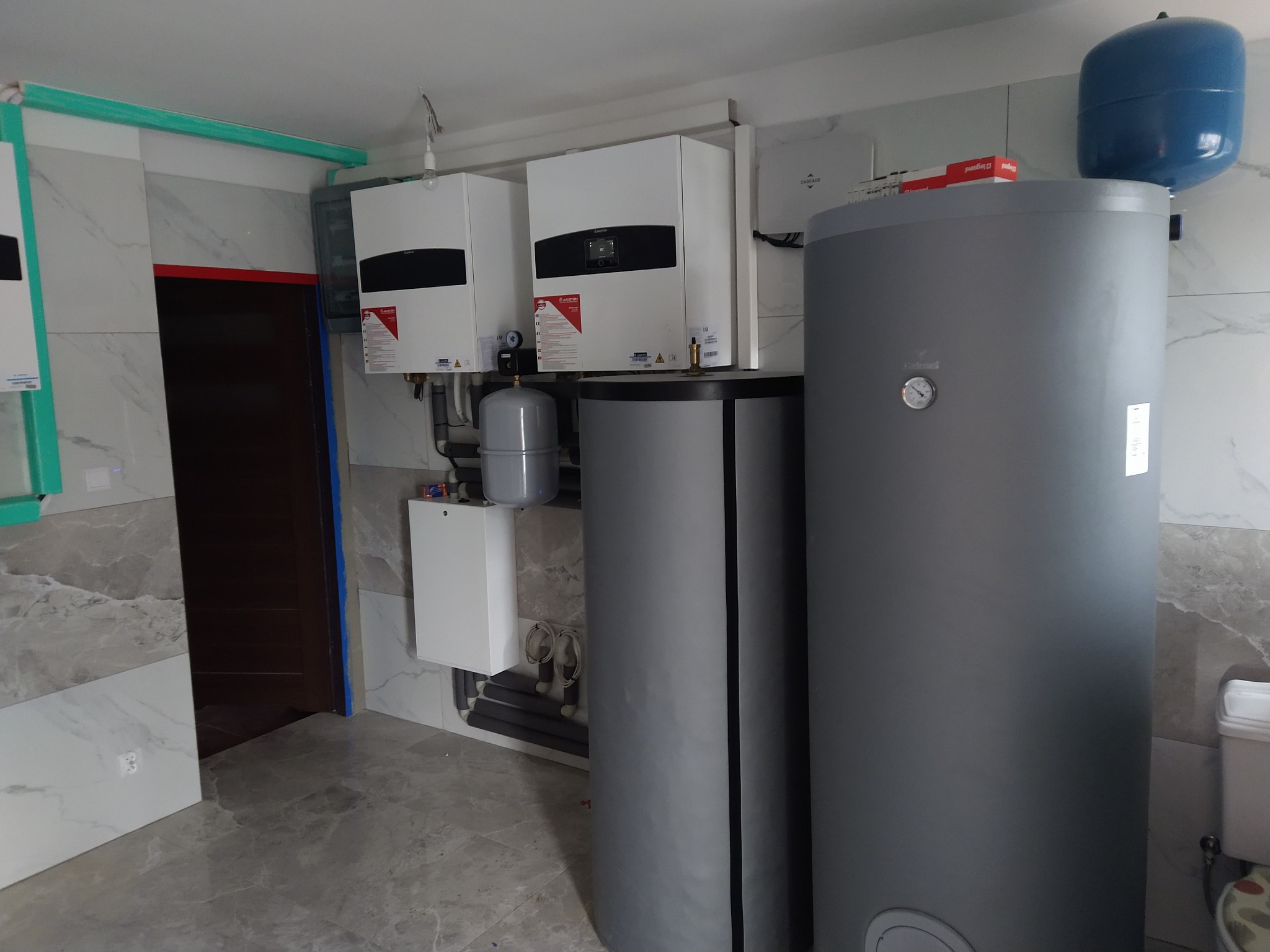 Dwa białe piece gazowe Ariston na ścianie z marmurowymi płytkami, obok szare zasobniki wody i niebieski zbiornik wyrównawczy. Widoczne rury i elementy instalacji.
