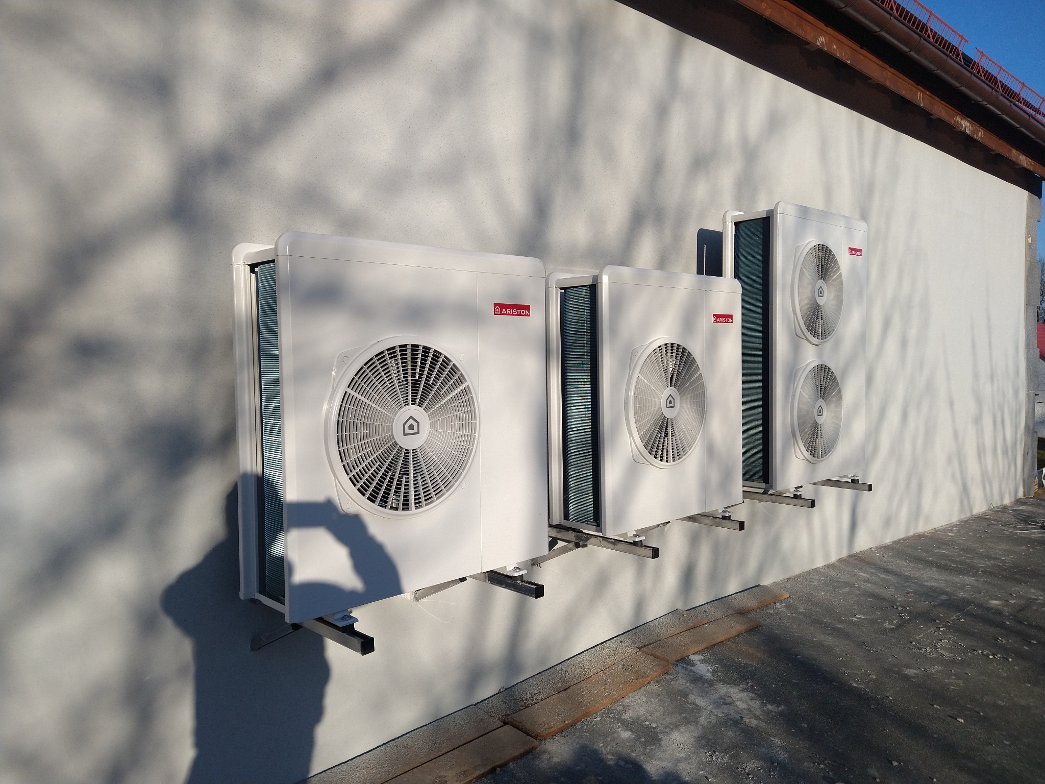 Trzy białe jednostki zewnętrzne klimatyzacji Ariston zamontowane na ścianie budynku, rzucające cień drzewa. Widoczne wsporniki montażowe i fragment dachu.