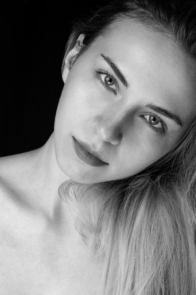 Portret młodej kobiety w czerni i bieli, z delikatnym makijażem i długimi włosami opadającymi na ramię, na ciemnym tle.