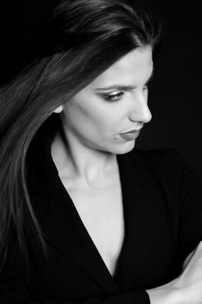 Czarno-białe, profilowe ujęcie kobiety w czarnej marynarce z rozpuszczonymi włosami, skierowanej w dół, z widocznym znamieniem na policzku.