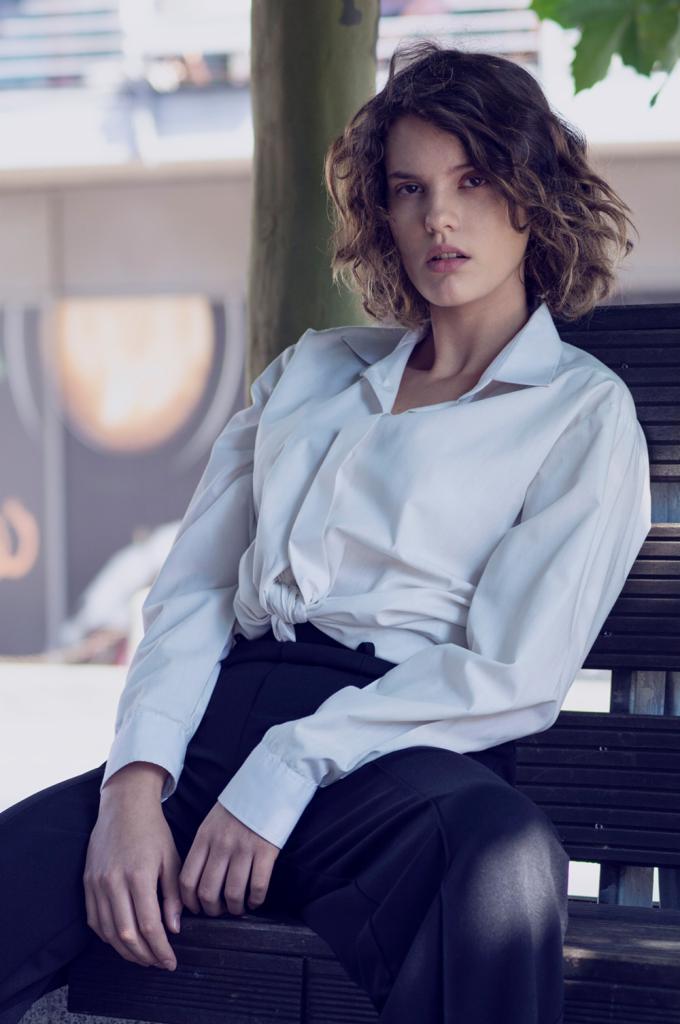 Portret kobiety w białej koszuli i czarnych spodniach, siedzącej na ławce w parku, z drzewem w tle i rozproszonym światłem.