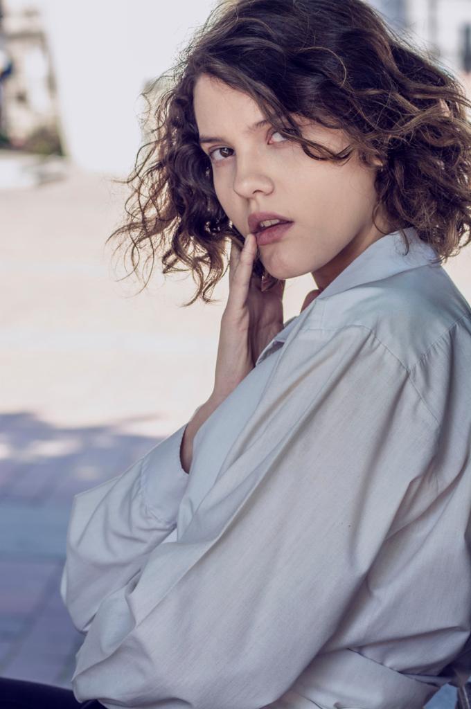 Portret młodej kobiety z kręconymi włosami, ubrana w białą koszulę, z palcem przy ustach, patrząca w bok.