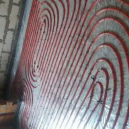 Układ czerwonych rur ogrzewania podłogowego na srebrnej folii izolacyjnej, widoczne mocowania i fragment surowego muru.