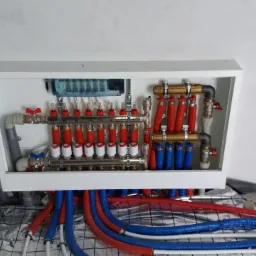 Rozdzielacz ogrzewania podłogowego w białej obudowie, widoczne czerwone i niebieskie rury, instalacja na folii aluminiowej z siatką.