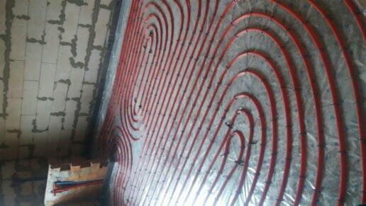 Układ czerwonych rur ogrzewania podłogowego na srebrnej folii izolacyjnej, widoczne mocowania i fragment surowego muru.