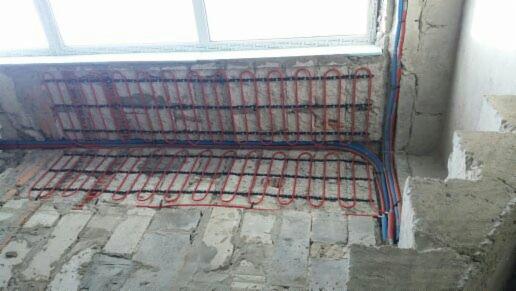 Montaż ogrzewania podłogowego w surowym stanie, widoczne pętle rurek grzewczych przymocowane do ściany z cegieł, obok pionowe rury instalacyjne oraz fragment schodów w stanie deweloperskim.