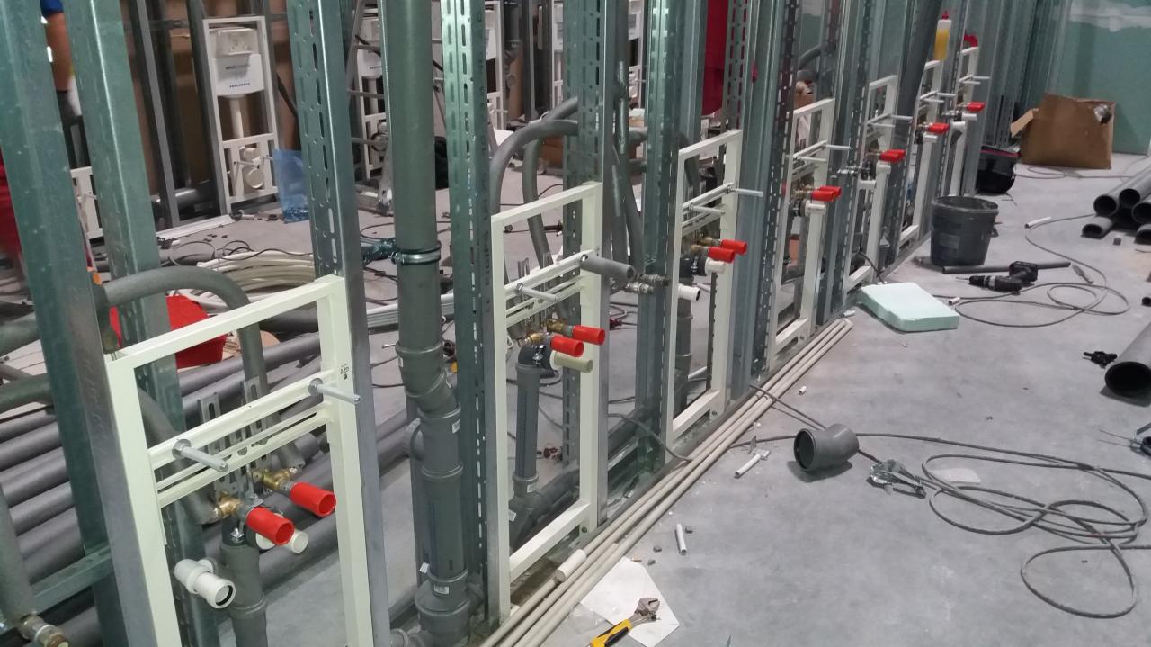 Wnętrze w trakcie prac instalacyjnych; widoczne szkielety ścian z zamontowanymi rurami, zaworami z czerwonymi uchwytami i elementami instalacji podtynkowej. Na podłodze leżą narzędzia i elementy...