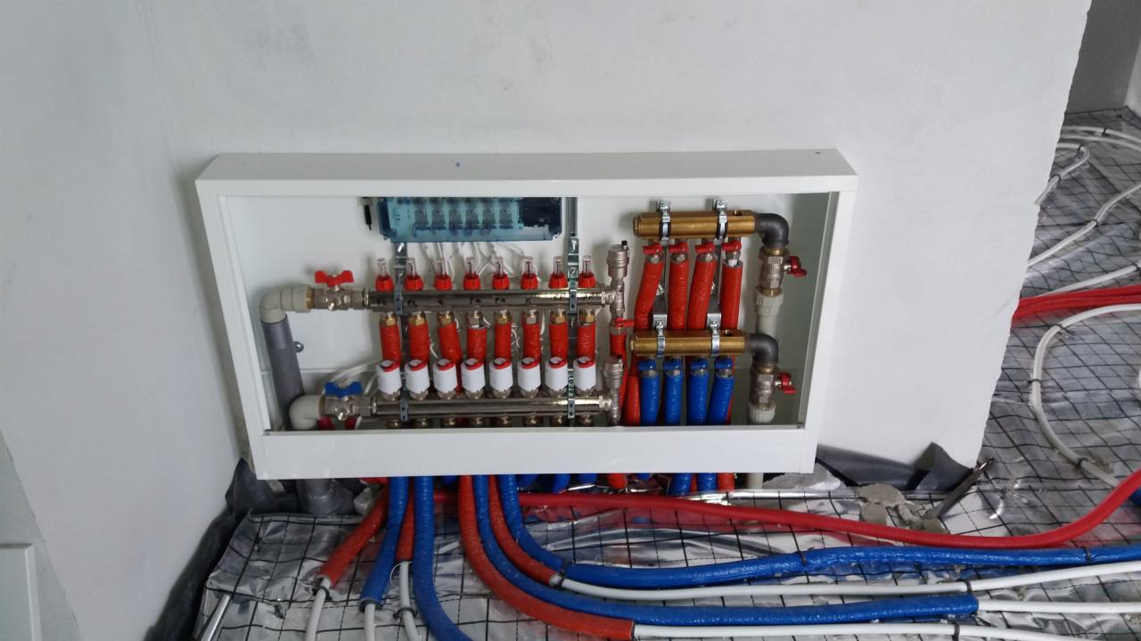 Rozdzielacz ogrzewania podłogowego w białej obudowie, widoczne czerwone i niebieskie rury, instalacja na folii aluminiowej z siatką.