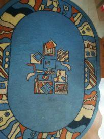 Owalny, błękitny dywan z geometrycznym wzorem w odcieniach brązu, beżu i pomarańczy. Widoczne ślady użytkowania i lekkie zabrudzenia na środku.