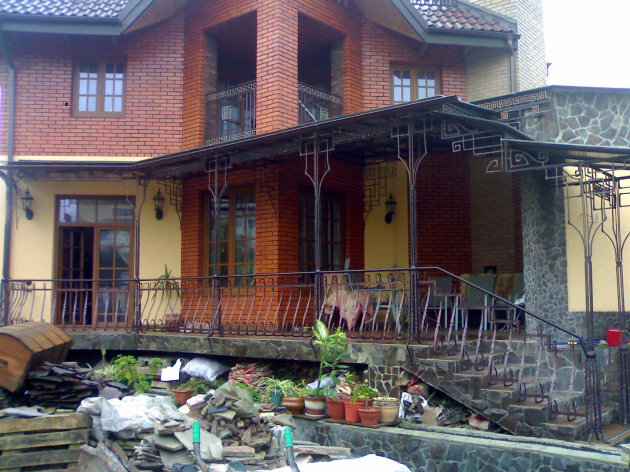 Kuta balustrada i zadaszenie nad tarasem domu z czerwonej cegły, widoczne fragmenty materiałów budowlanych.