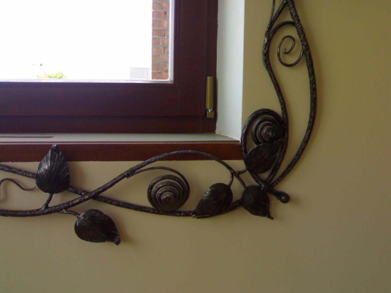 Kuta, metalowa dekoracja okienna z motywem roślinnym, zamontowana na beżowej ścianie, widoczna część okna z brązową ramą.