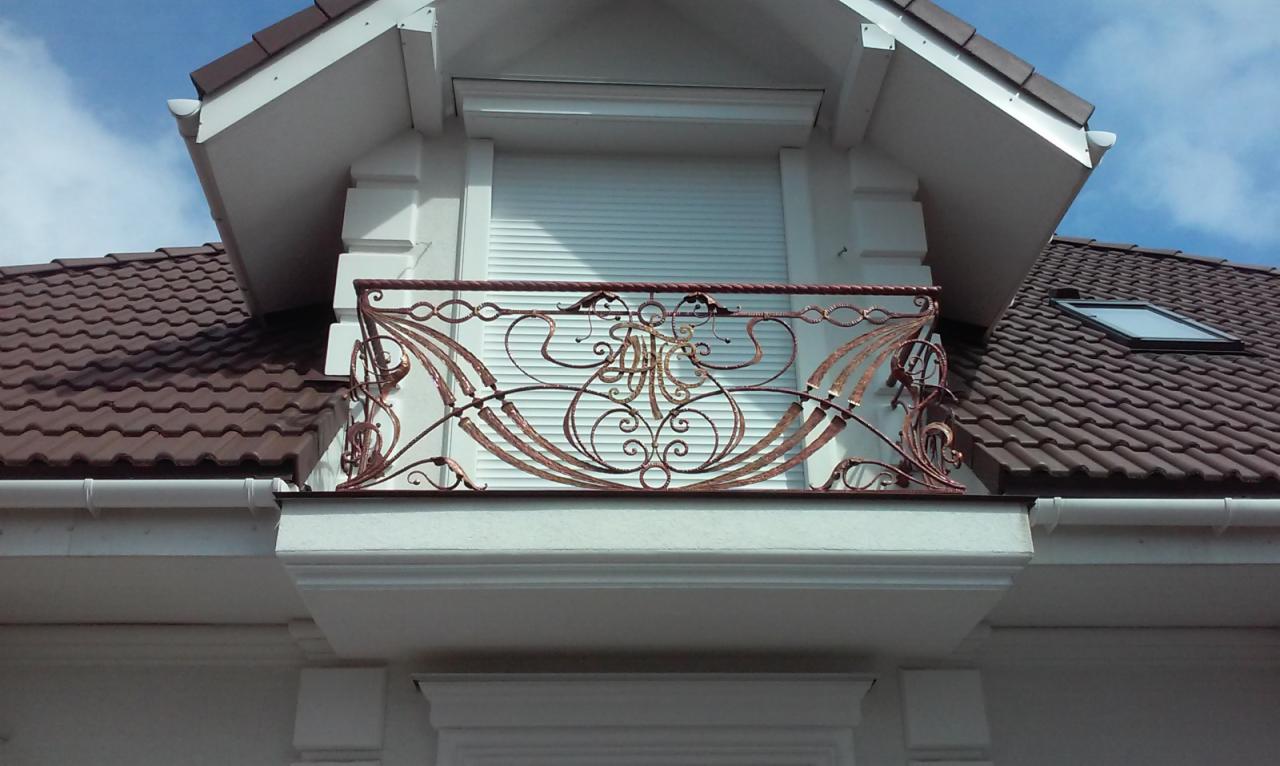 Dekoracyjna, kuta balustrada balkonowa w kolorze miedzianym, z ozdobnym motywem roślinnym i inicjałami, na tle białej fasady budynku z zamkniętą roletą okienną i brązowym dachem.