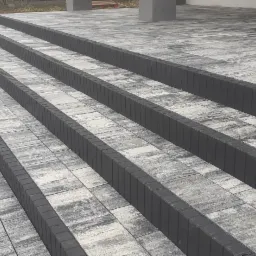 Schody wejściowe wyłożone szarą kostką brukową z ciemnoszarymi obrzeżami, widoczne z boku pod kątem, z fragmentem trawnika w tle.