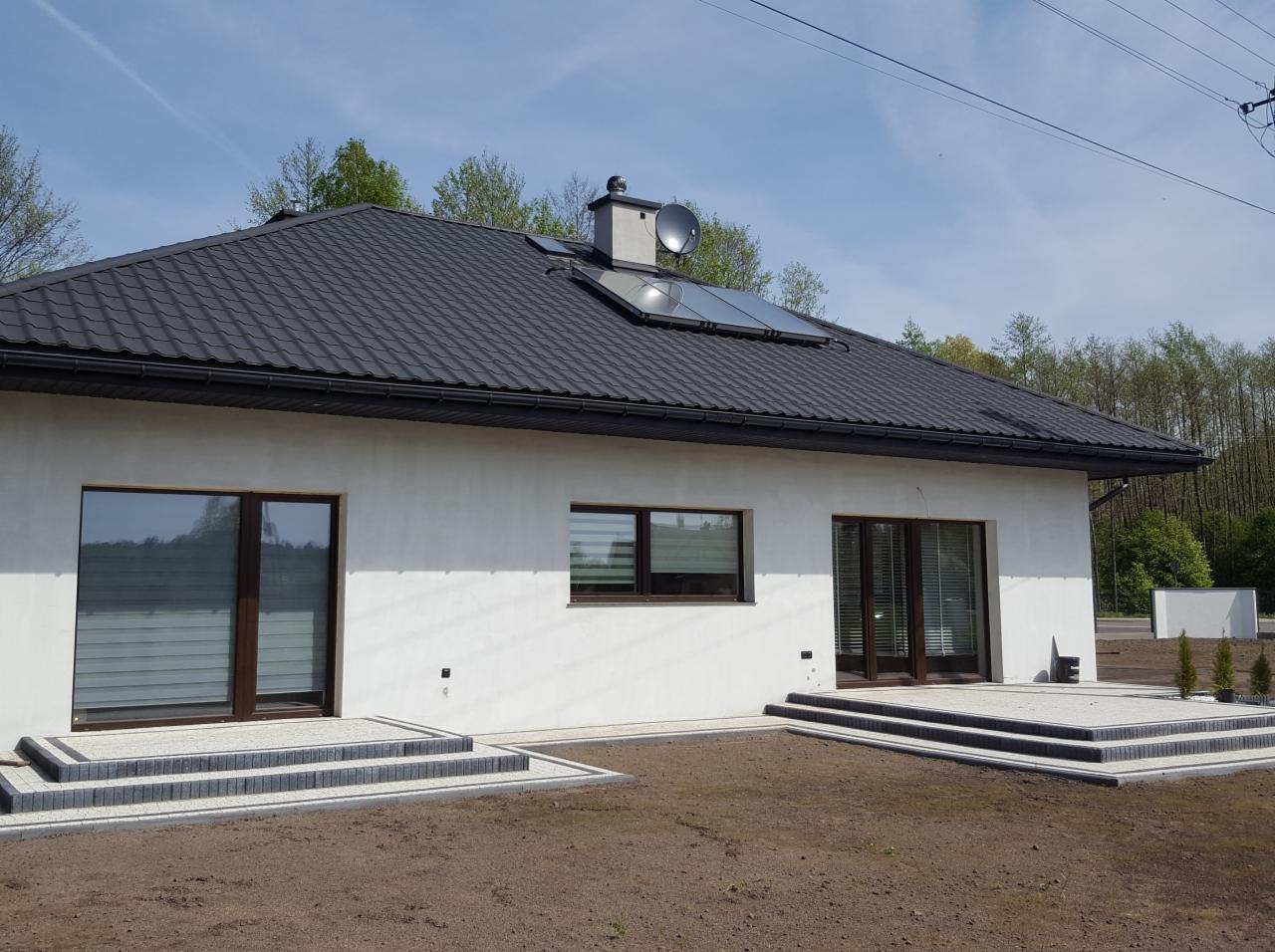 Nowoczesny dom jednorodzinny z antracytowym dachem, panelami słonecznymi i kostką brukową tworzącą schody wejściowe i taras.