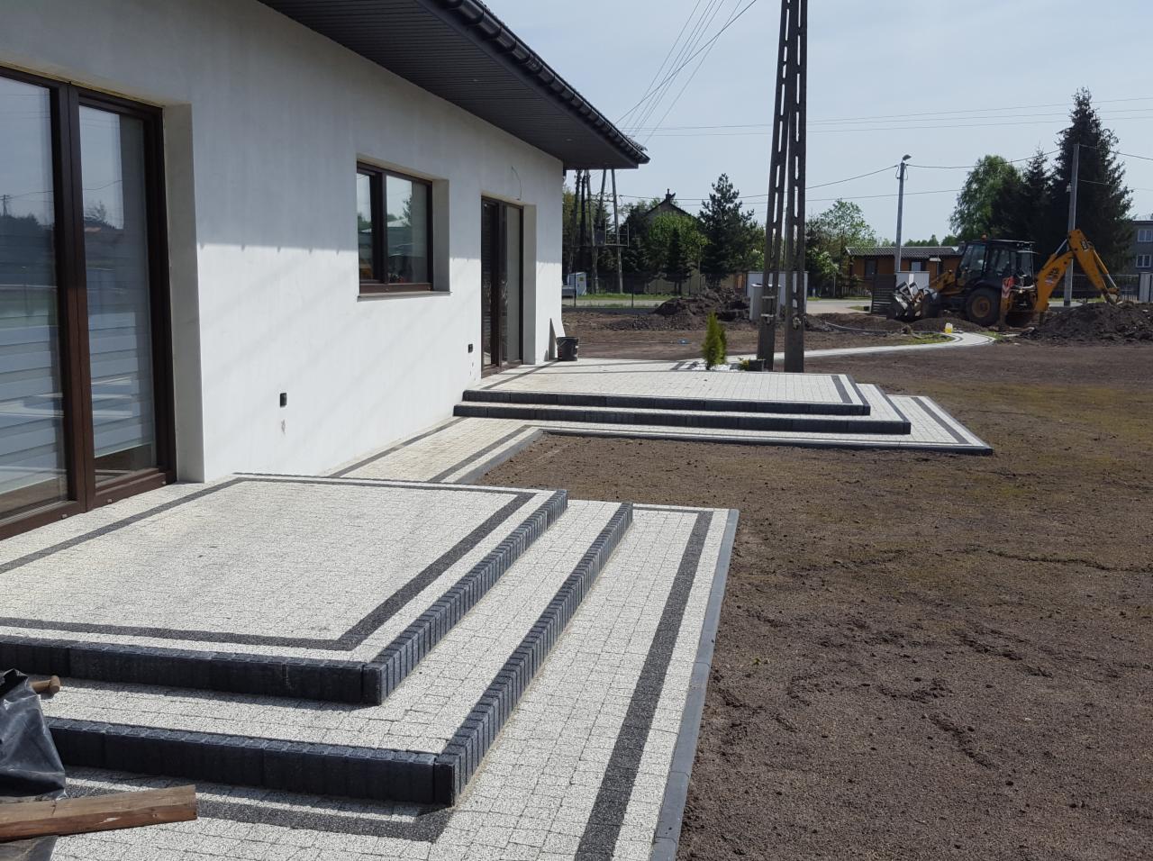 Schody i taras wyłożone jasnoszarą kostką z ciemnoszarym obramowaniem, widok na nowo obsianą trawę i koparkę w tle.