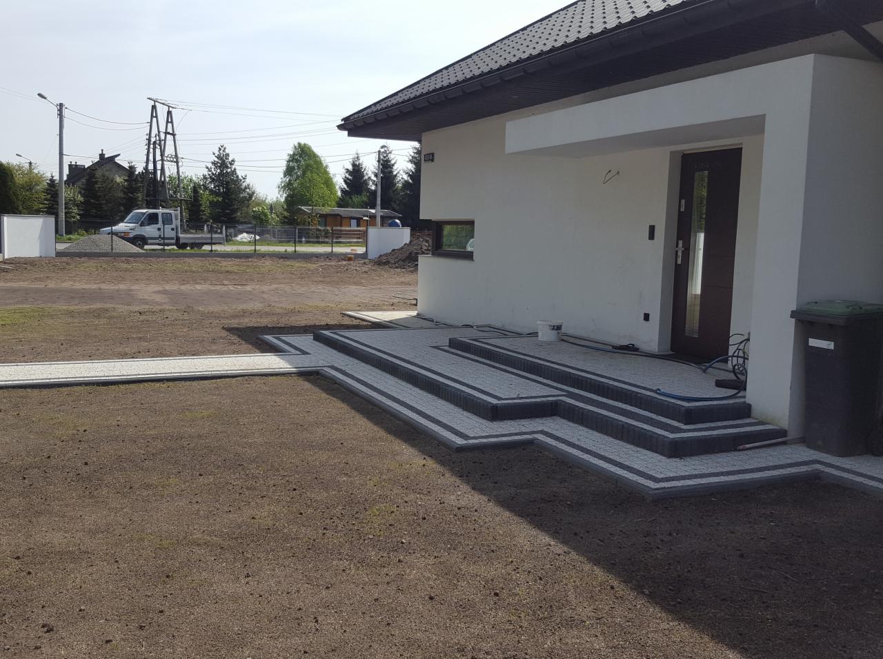 Nowoczesny dom z wejściem wyłożonym szarą kostką brukową z ciemnymi obrzeżami, widoczne schody i ścieżka prowadząca do trawnika z przygotowaną ziemią pod zasiew.