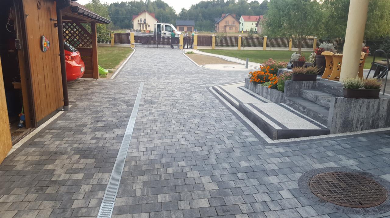 Ułożona szara kostka brukowa na podjeździe z odpływem liniowym, widoczne schody z kostki granitowej prowadzące do domu z rabatami kwiatowymi i drewniany garaż z czerwonym samochodem w tle.