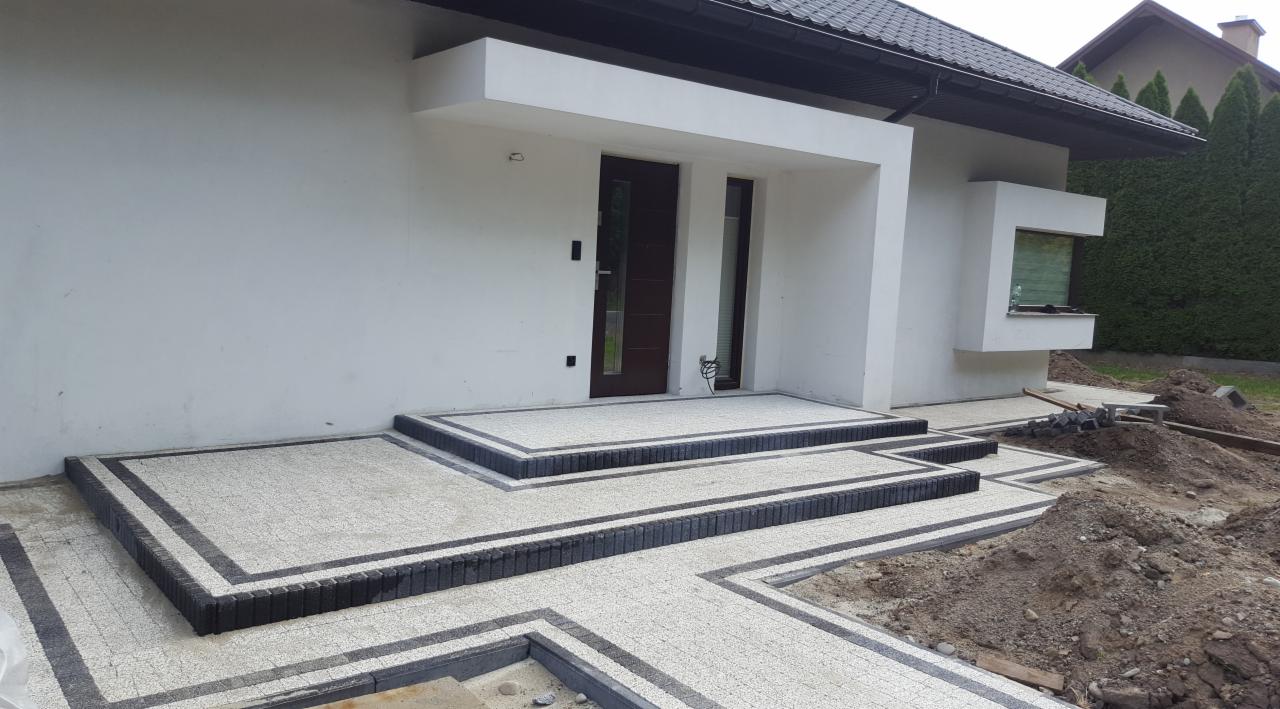 Wykończone schody wejściowe do nowoczesnego domu z jasnej kostki brukowej z ciemną obwódką, widoczne narzędzia i ziemia obok ścieżki.