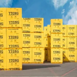 Żółte bloczki Ytong ułożone w stosy na zewnątrz, pod błękitnym niebem z chmurami. Widoczna tekstura bloczków i logotyp Ytong. Perspektywa z dołu.