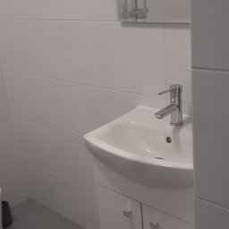 PHU SZAFIR - Biała umywalka z szafką w wyremontowanej łazience, widok na baterię i lustro z kabiną prysznicową. Szare płytki podłogowe, minimalistyczny styl.