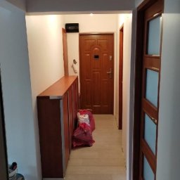 PHU SZAFIR - Wykończony korytarz z drewnianą szafką, drzwiami i panelami podłogowymi w odcieniach beżu. Widoczny worek z materiałami budowlanymi i wiadro z farbą.