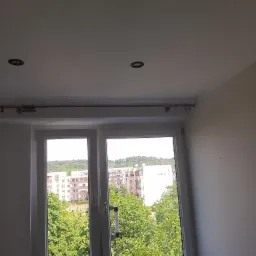 Pok&oacute;j po remoncie z widokiem na zieleń przez okno; widoczne białe ściany, sufit z dwoma punktowymi lampami, okno z termometrem i karnisz. Na parapecie leżą dokumenty.