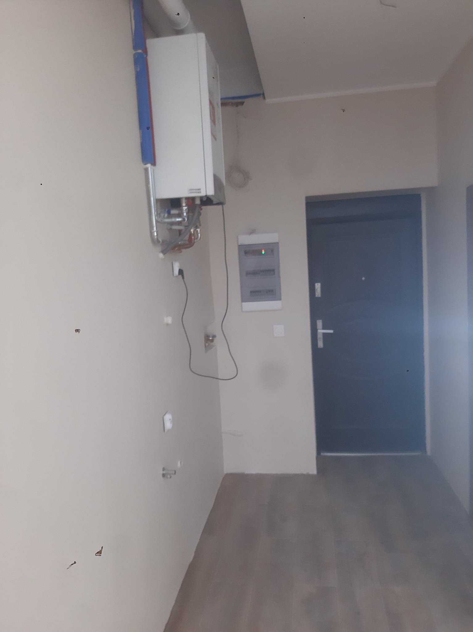 Wnętrze mieszkania w trakcie prac instalacyjnych: zamontowany piec gazowy na ścianie, widoczna instalacja wodna i elektryczna, skrzynka elektryczna, drzwi wejściowe.