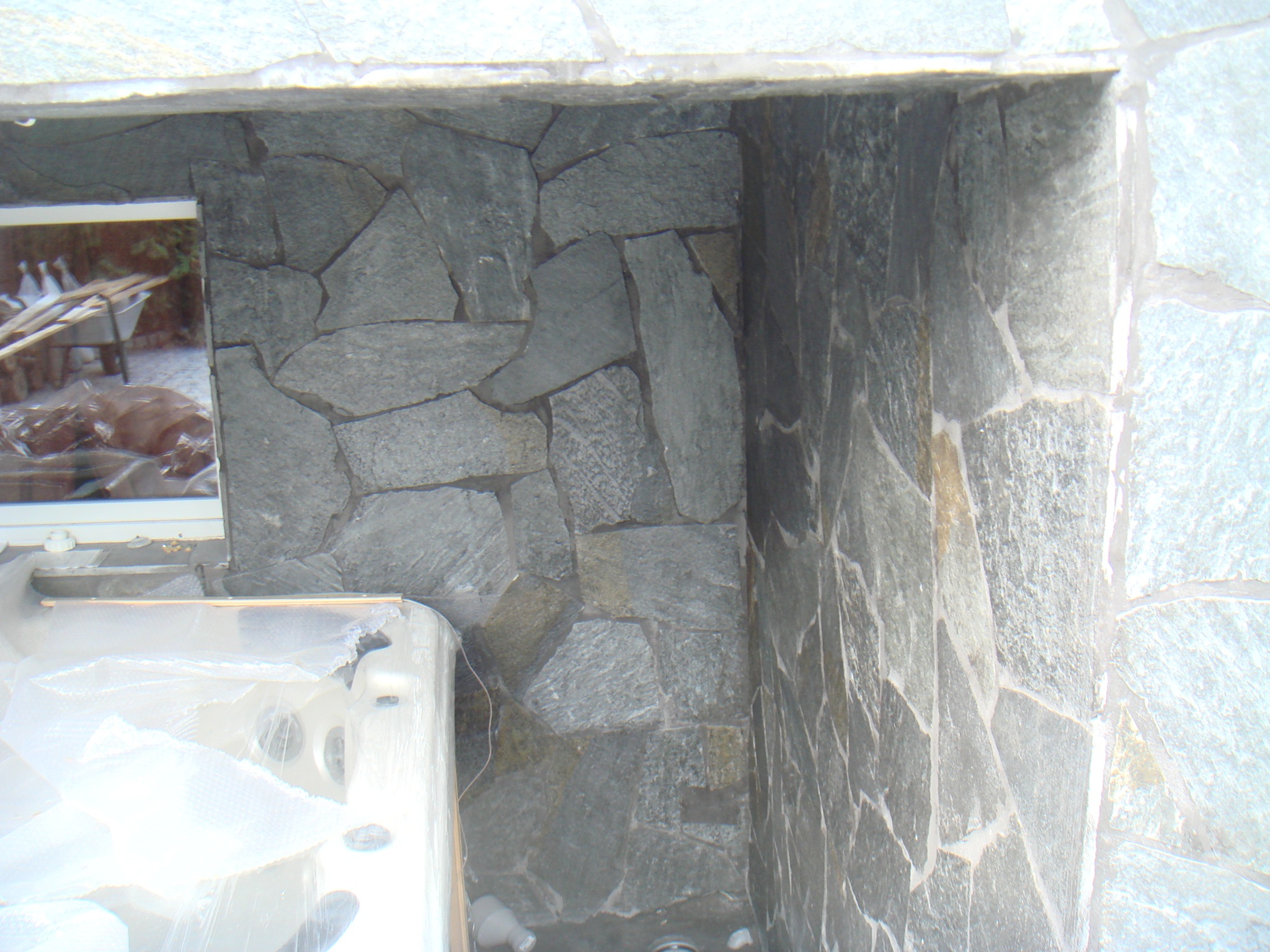 Fragment ściany wyłożonej nieregularnymi płytami kamiennymi w odcieniach szarości, z widoczną wnęką i fragmentem wanny jacuzzi w folii ochronnej.