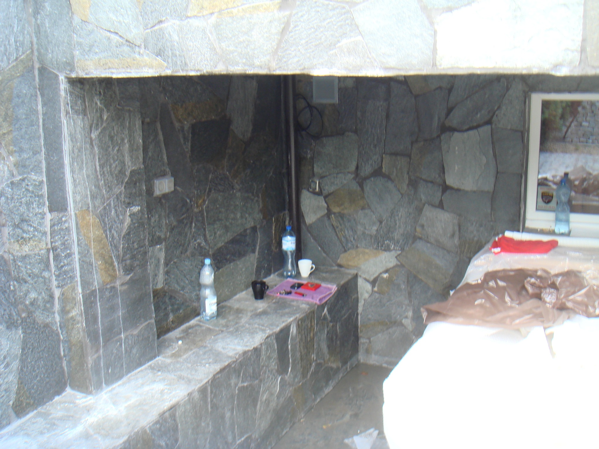 Ściana wyłożona nieregularnymi kamiennymi płytami, z półką na której stoją butelki z wodą, kubki i inne przedmioty; widoczne również fragmenty zakrytego mebla lub urządzenia.