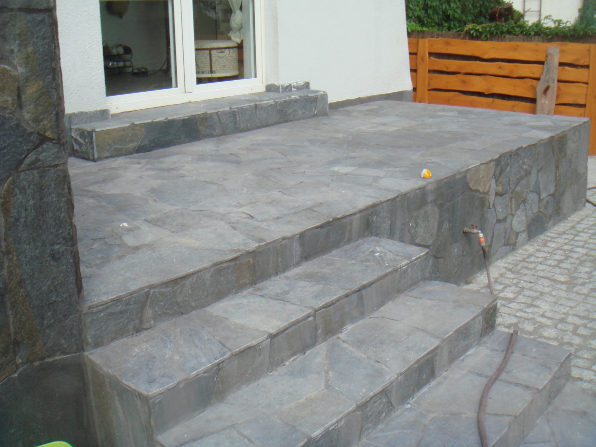 Kamienne schody prowadzące do tarasu wyłożonego nieregularnymi płytami, z widocznym kranem i wężem ogrodowym oraz fragmentem drewnianego ogrodzenia w tle.