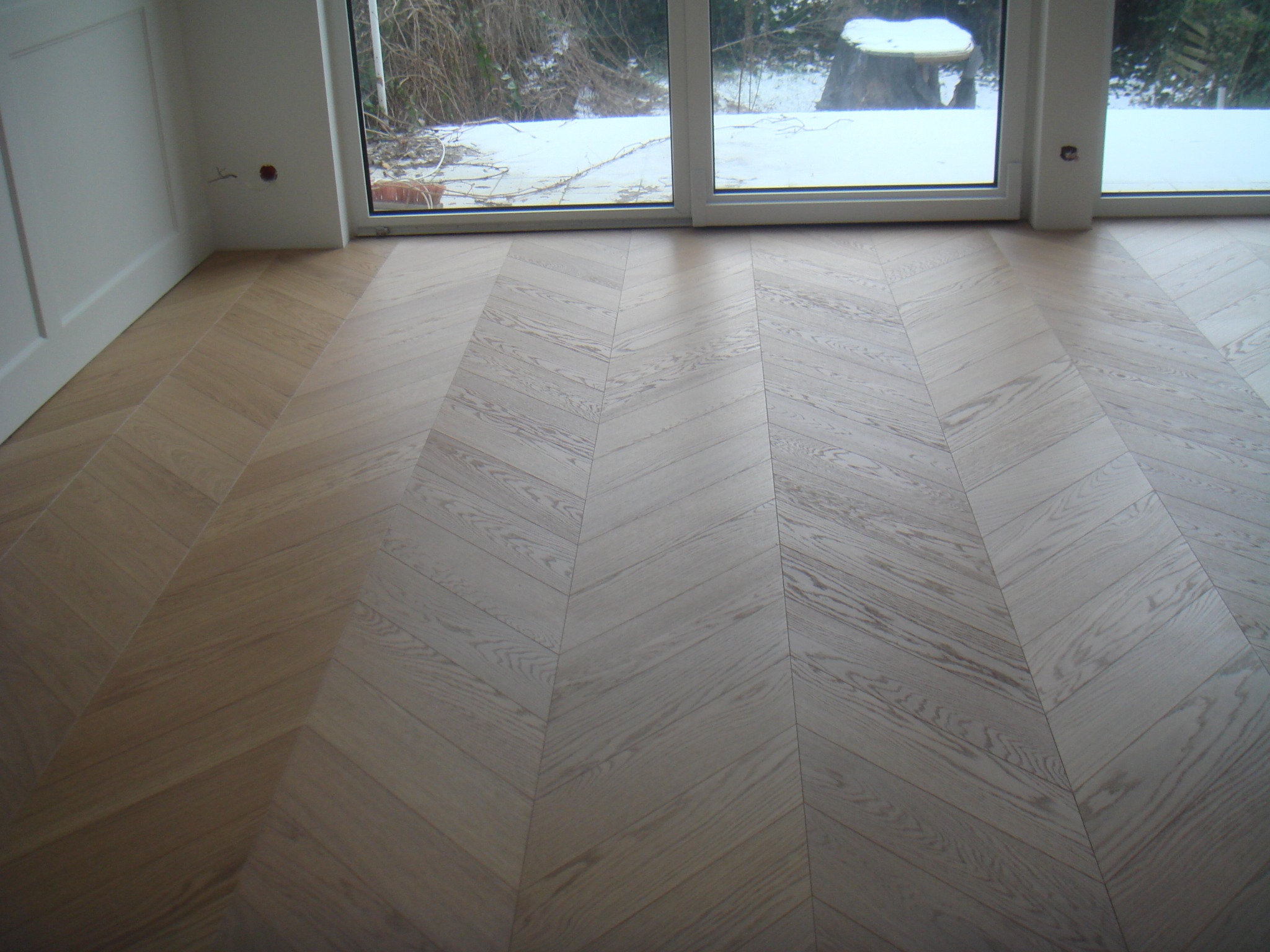 Ułożona jodełka z jasnych paneli podłogowych, widok na okno z zimowym krajobrazem, widoczne puszki instalacyjne w ścianie.