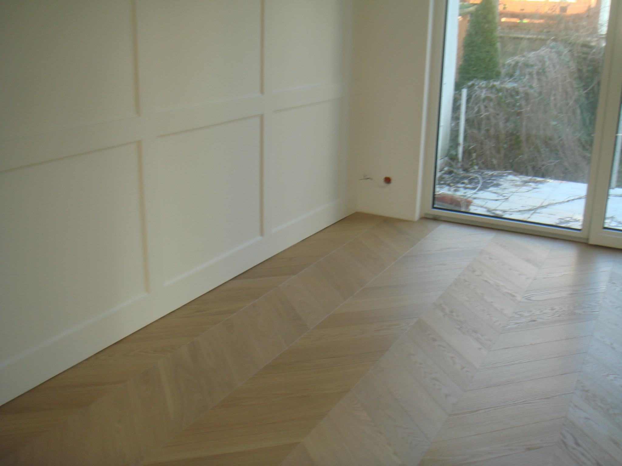 Jasny parkiet ułożony we wzór jodełki francuskiej w pokoju z białymi ścianami z dekoracyjnymi panelami i dużym oknem balkonowym z widokiem na ogród.