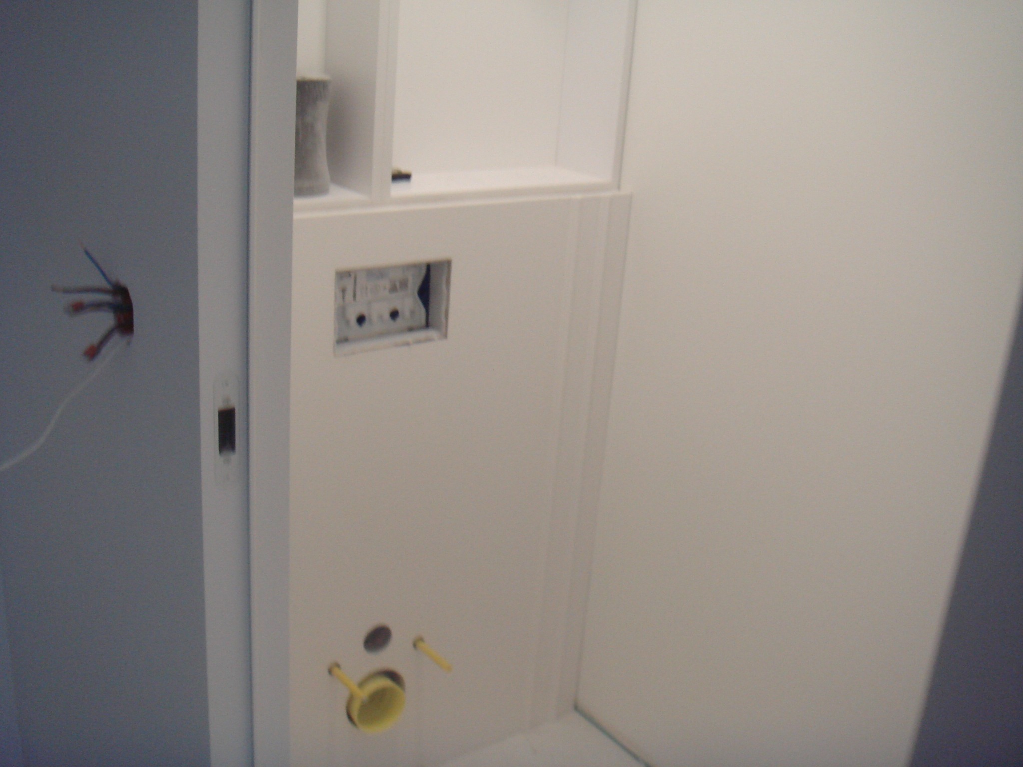 Biała ściana z instalacją podtynkową WC w trakcie montażu, widoczne otwory na rury i puszkę elektryczną, fragment instalacji elektrycznej wystaje ze ściany.