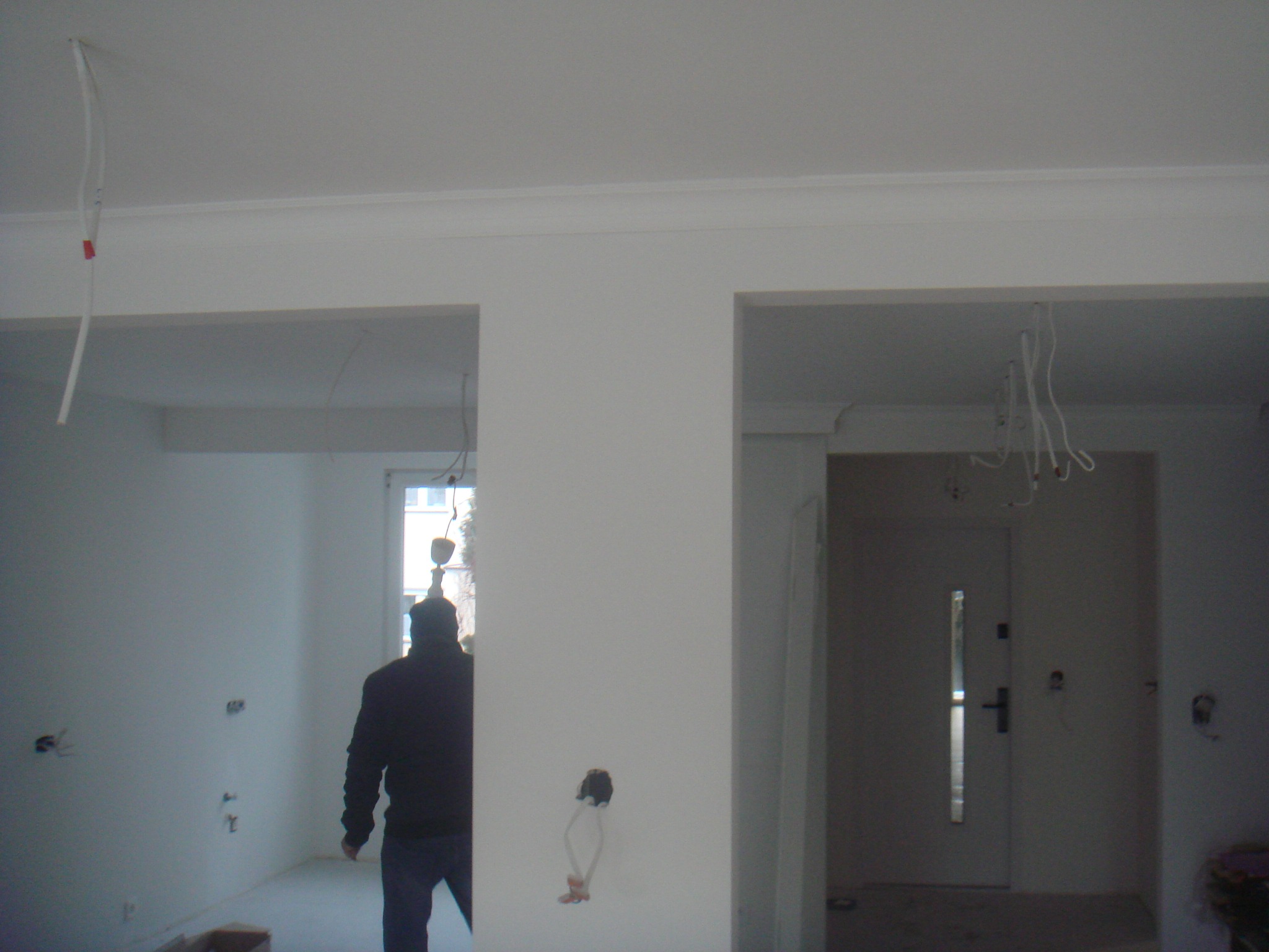 Wnętrze w trakcie prac wykończeniowych: widoczne kable wystające ze ściany i sufitu, otwory w ścianach, białe ściany z listwami dekoracyjnymi pod sufitem, sylwetka osoby w oddali.