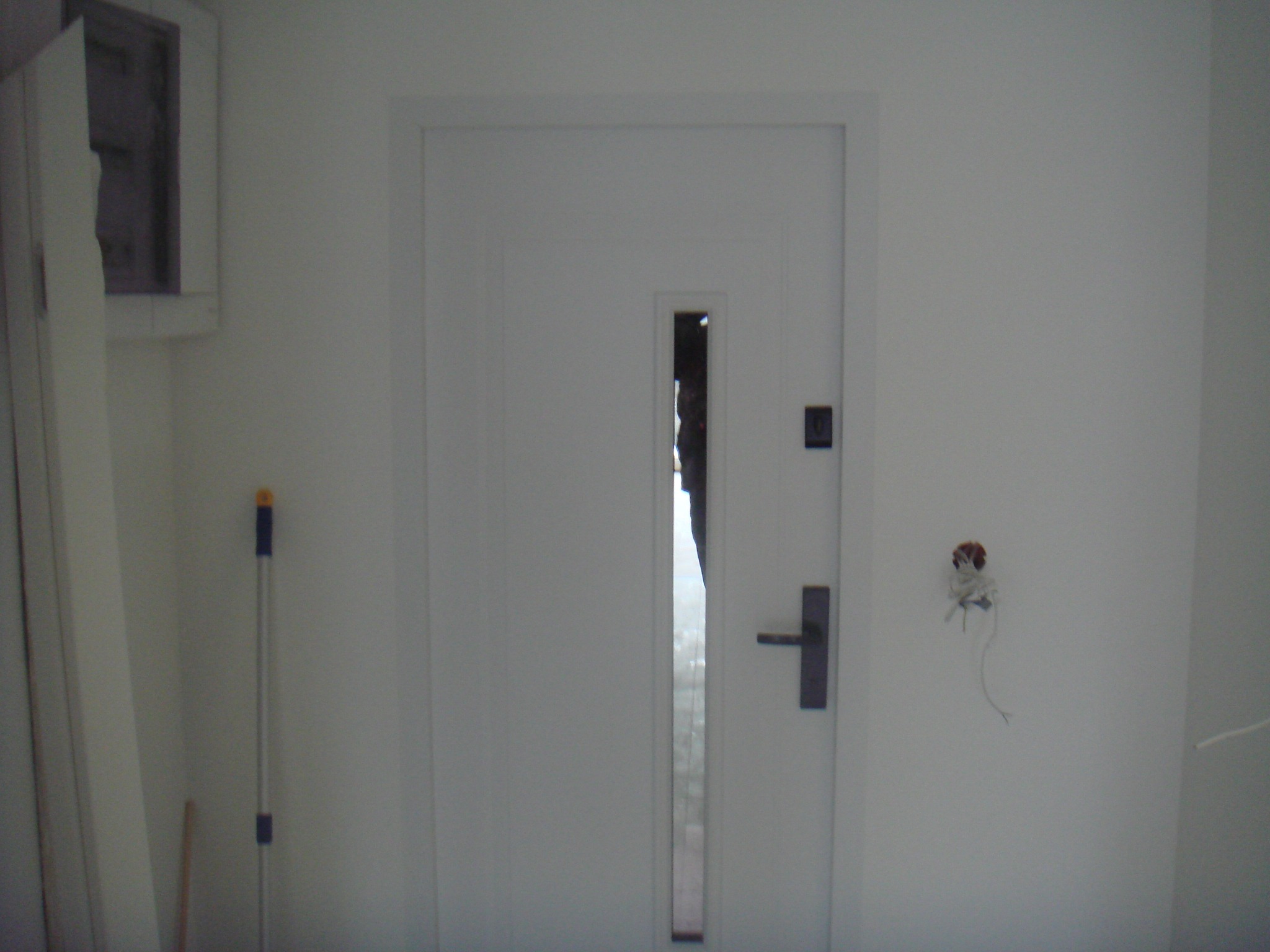 Białe drzwi wewnętrzne z wąskim przeszkleniem pionowym, widoczny fragment ściany z wystającymi kablami elektrycznymi i listwa przypodłogowa oparta o ścianę.