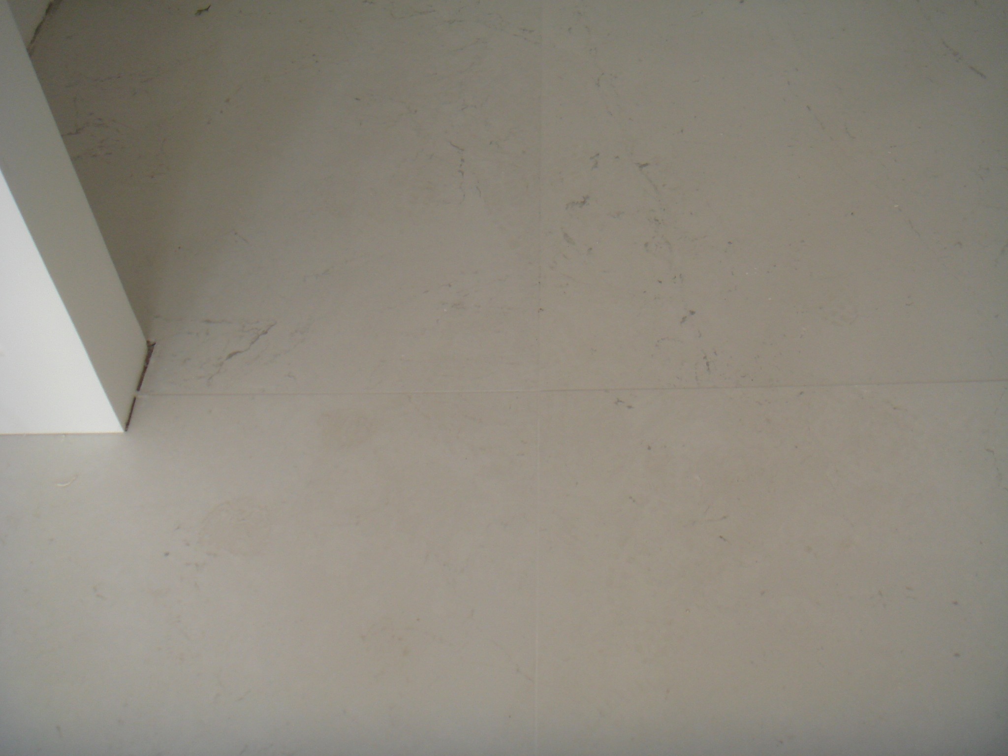 Jasnoszara podłoga wyłożona płytkami ceramicznymi z delikatnym wzorem, widoczny fragment białej ściany w lewym górnym rogu.