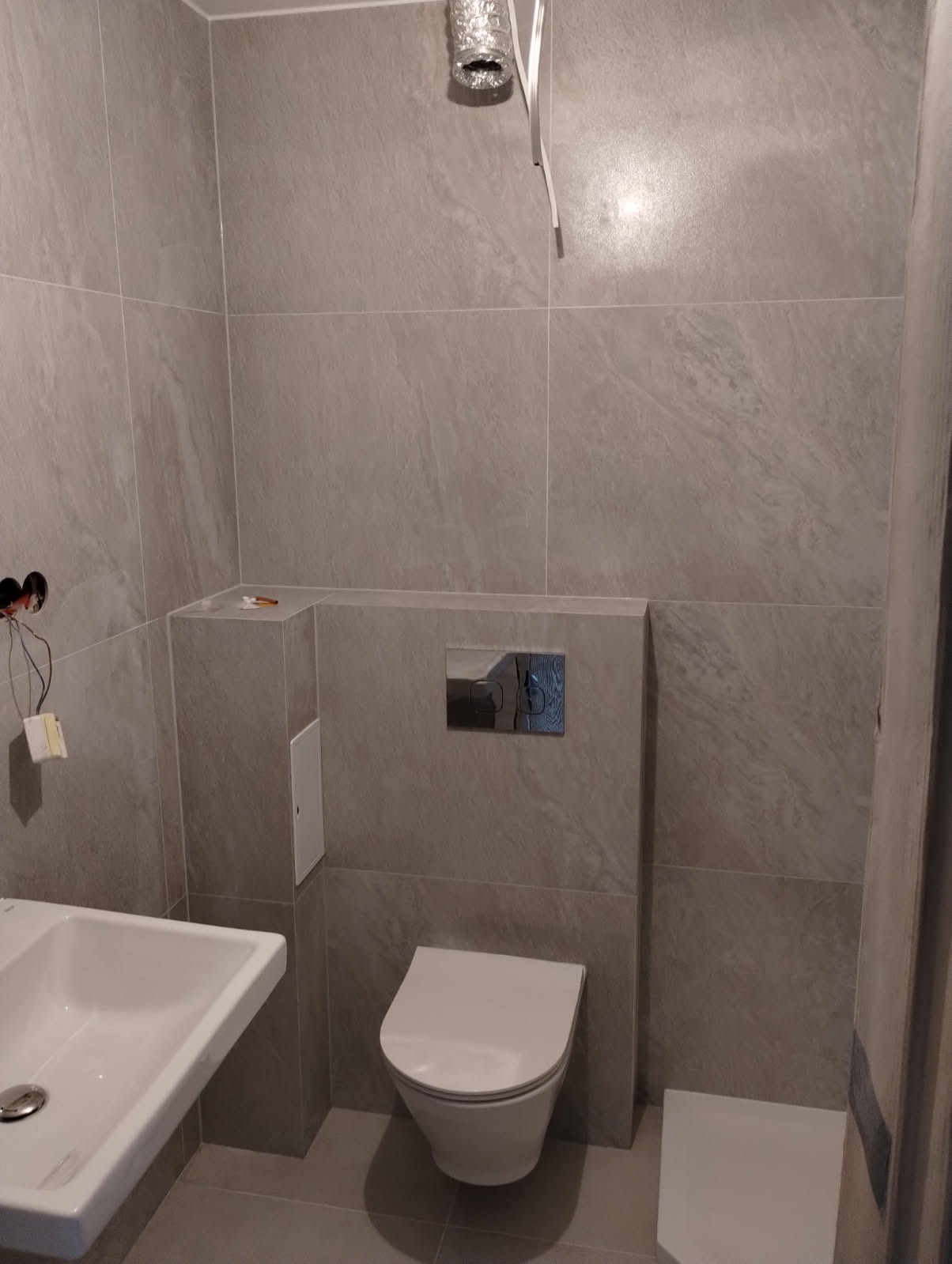 Wykończona łazienka w szarościach: umywalka, wisząca toaleta, płytki imitujące kamień. Widoczne niedokończone podłączenie wentylacji i instalacji elektrycznej.