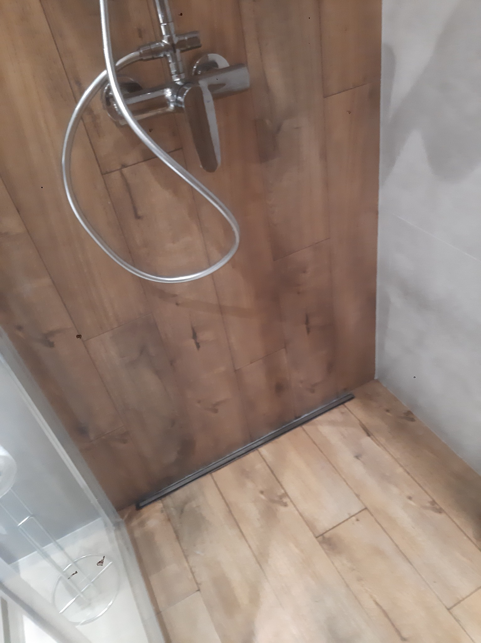 Kabina prysznicowa z panelami imitującymi drewno na ścianach i podłodze, widoczny chromowany kran i odpływ liniowy.