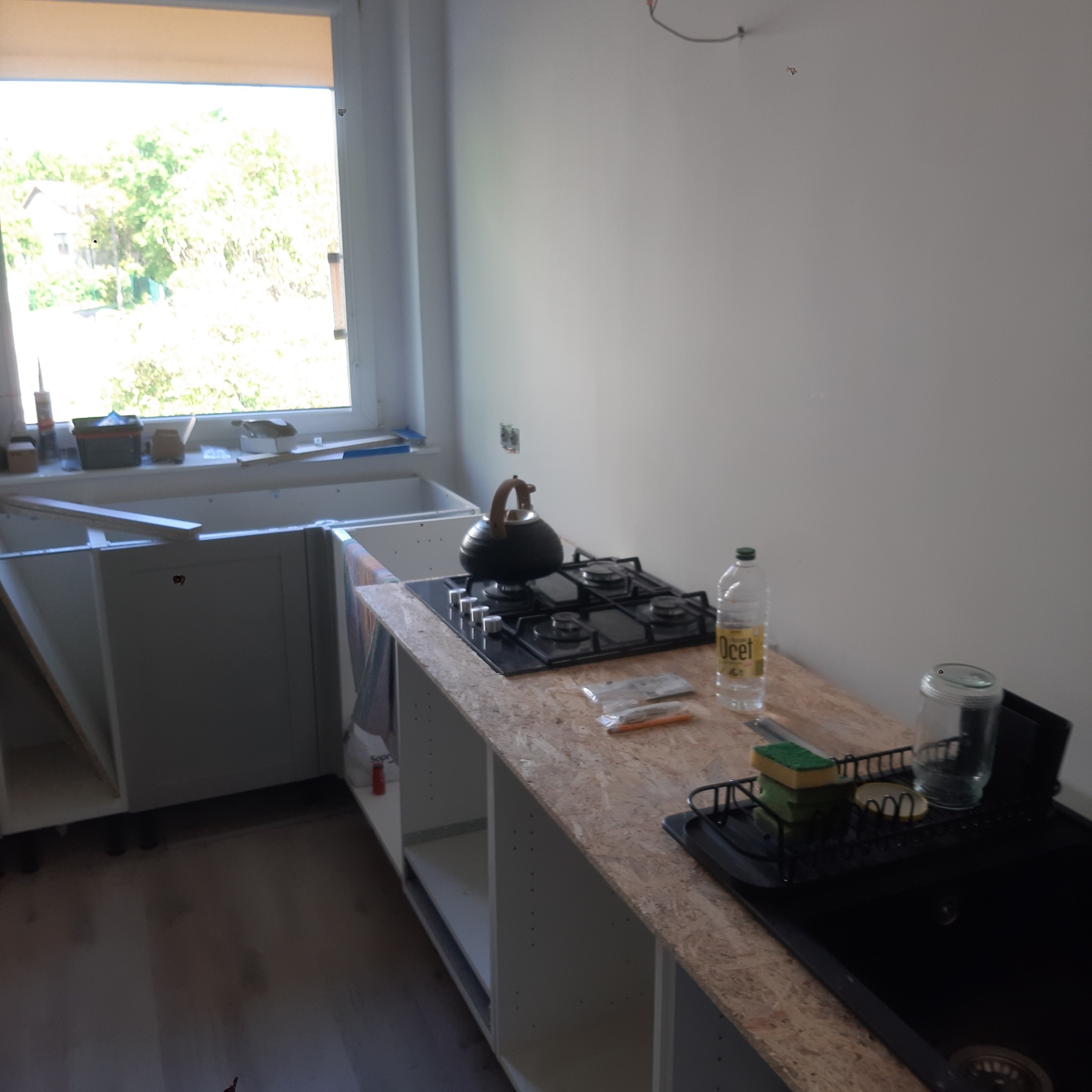 Montaż mebli kuchennych: surowe blaty z płyty OSB,  częściowo zamontowane szafki, płyta gazowa z czajnikiem, okno w tle. Wnętrze w trakcie prac wykończeniowych.