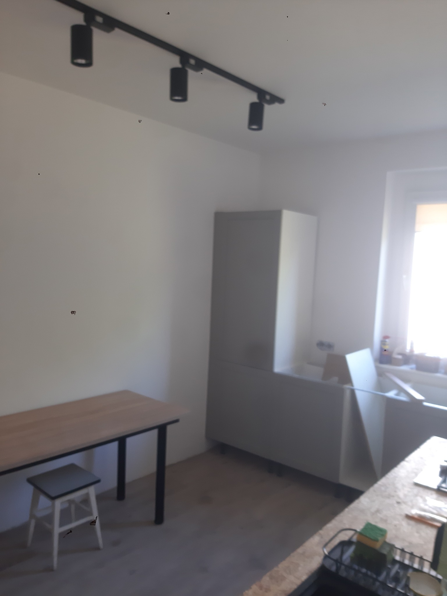 Wnętrze kuchni w trakcie montażu: widoczne szafki, stół z krzesłem, oświetlenie sufitowe. Elementy wymagają jeszcze wykończenia, co wskazuje na etap prac.