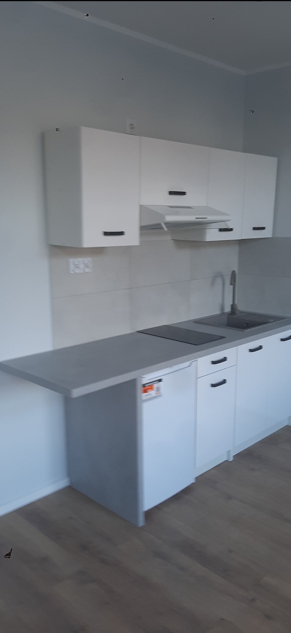 Nowoczesna, biała zabudowa kuchenna z szarym blatem i płytkami. Minimalistyczny design, czarne uchwyty, płyta indukcyjna i zlew. Widok na fragment podłogi.