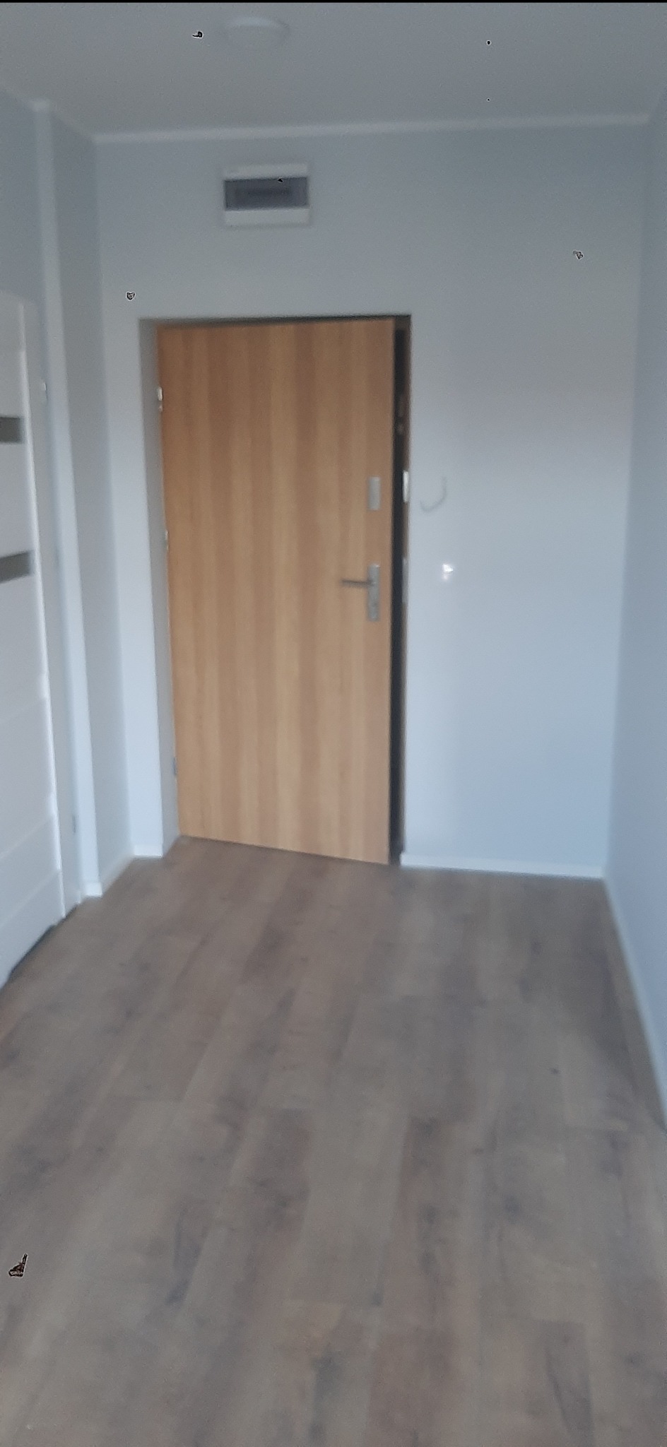 Jasne panele podłogowe w korytarzu z drewnianymi drzwiami i białymi ścianami. Widoczny fragment białych drzwi po lewej stronie.